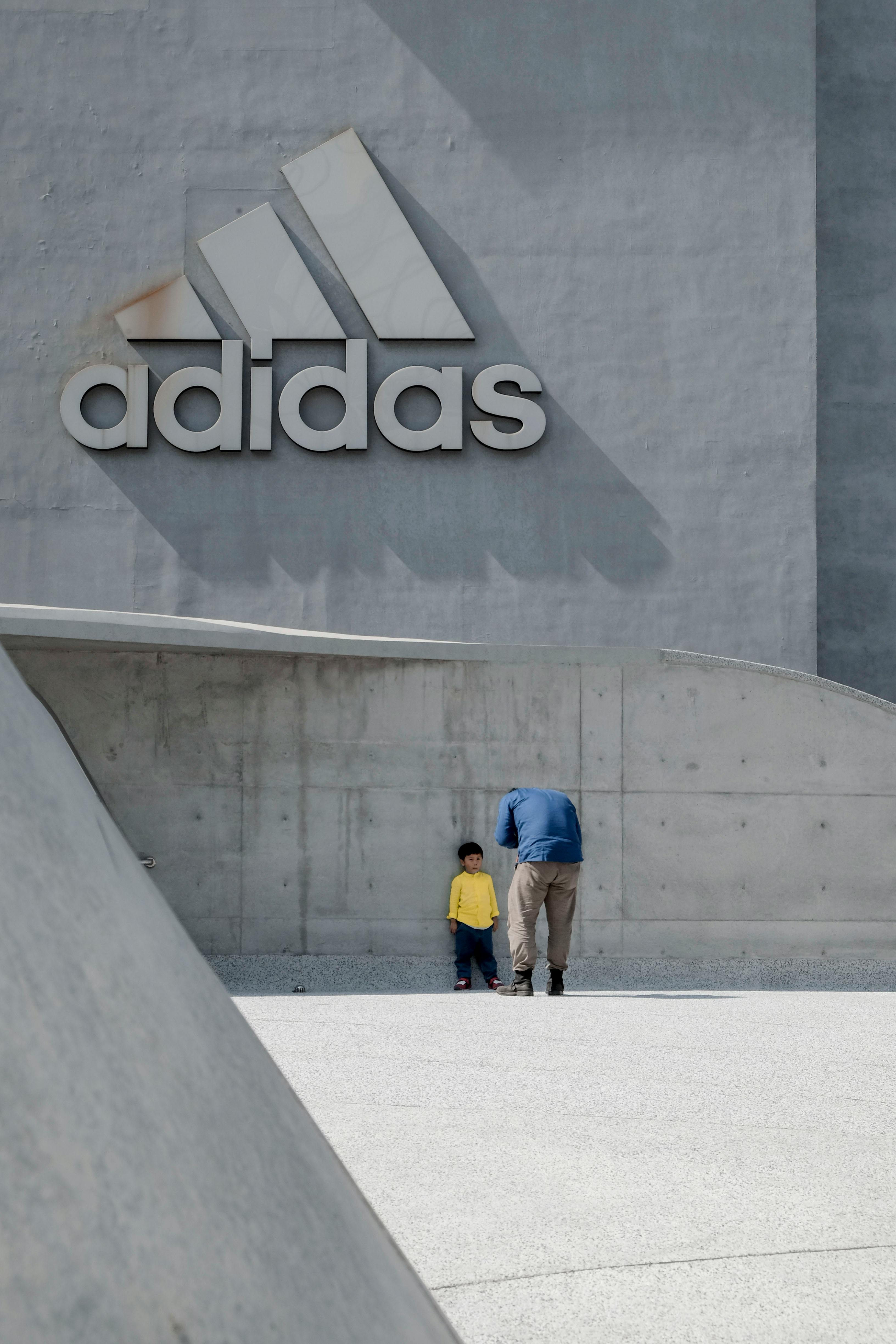 A father and child stand by a large Adidas logo on a modern building facade.