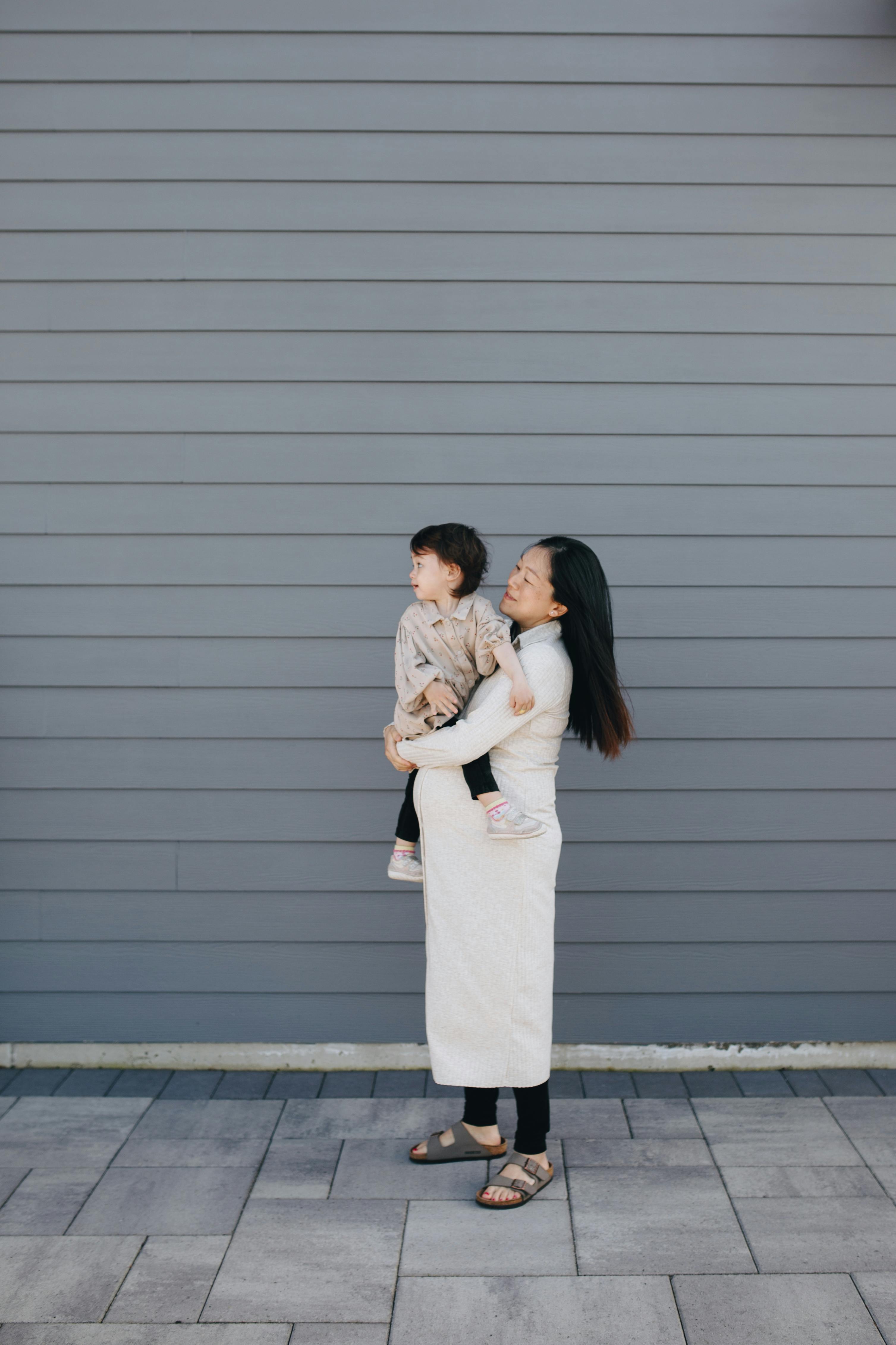 A mother joyfully holds her young daughter outdoors against a modern grey wall.