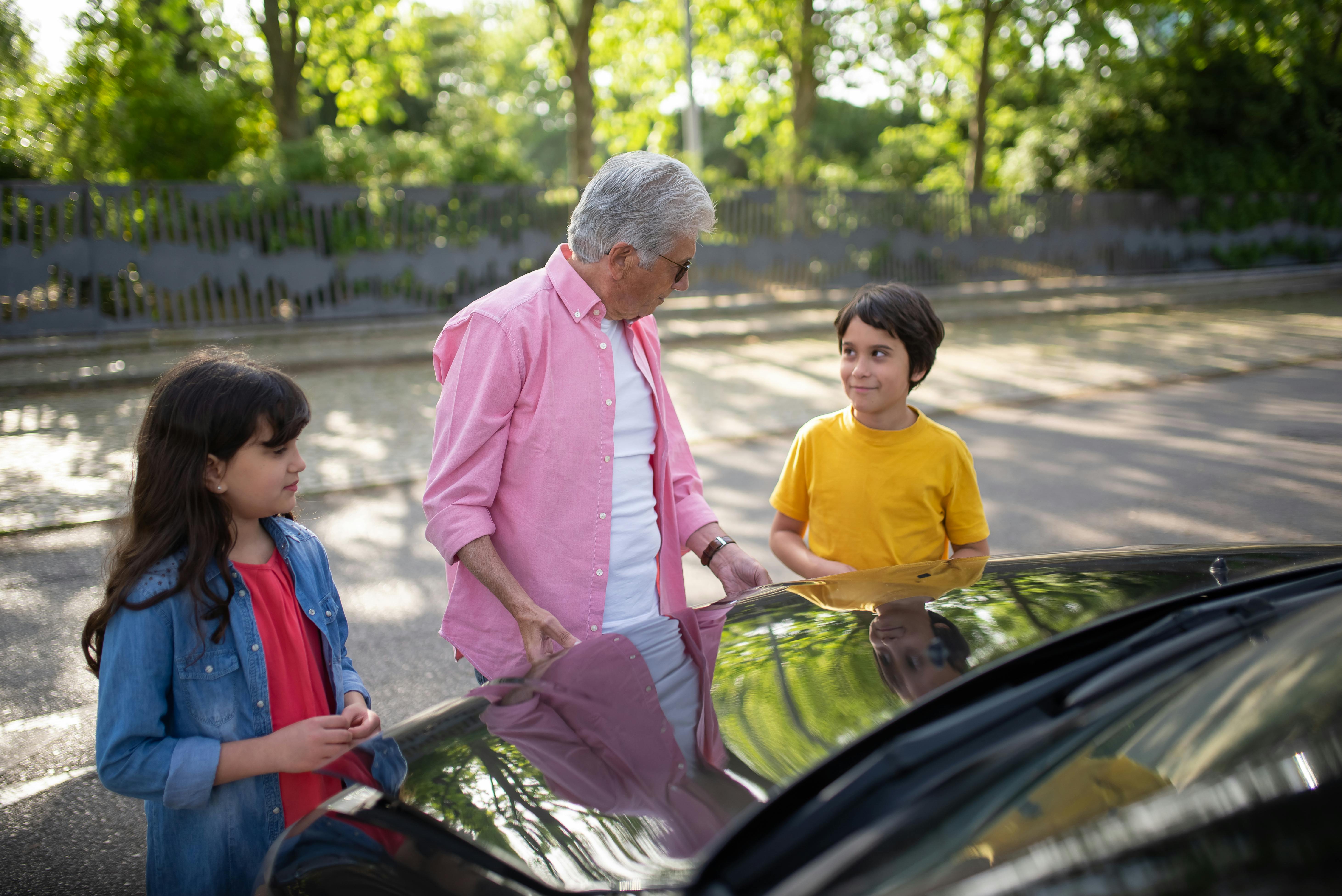 A grandfather spends time with his grandchildren by a car on a sunny day outdoors.