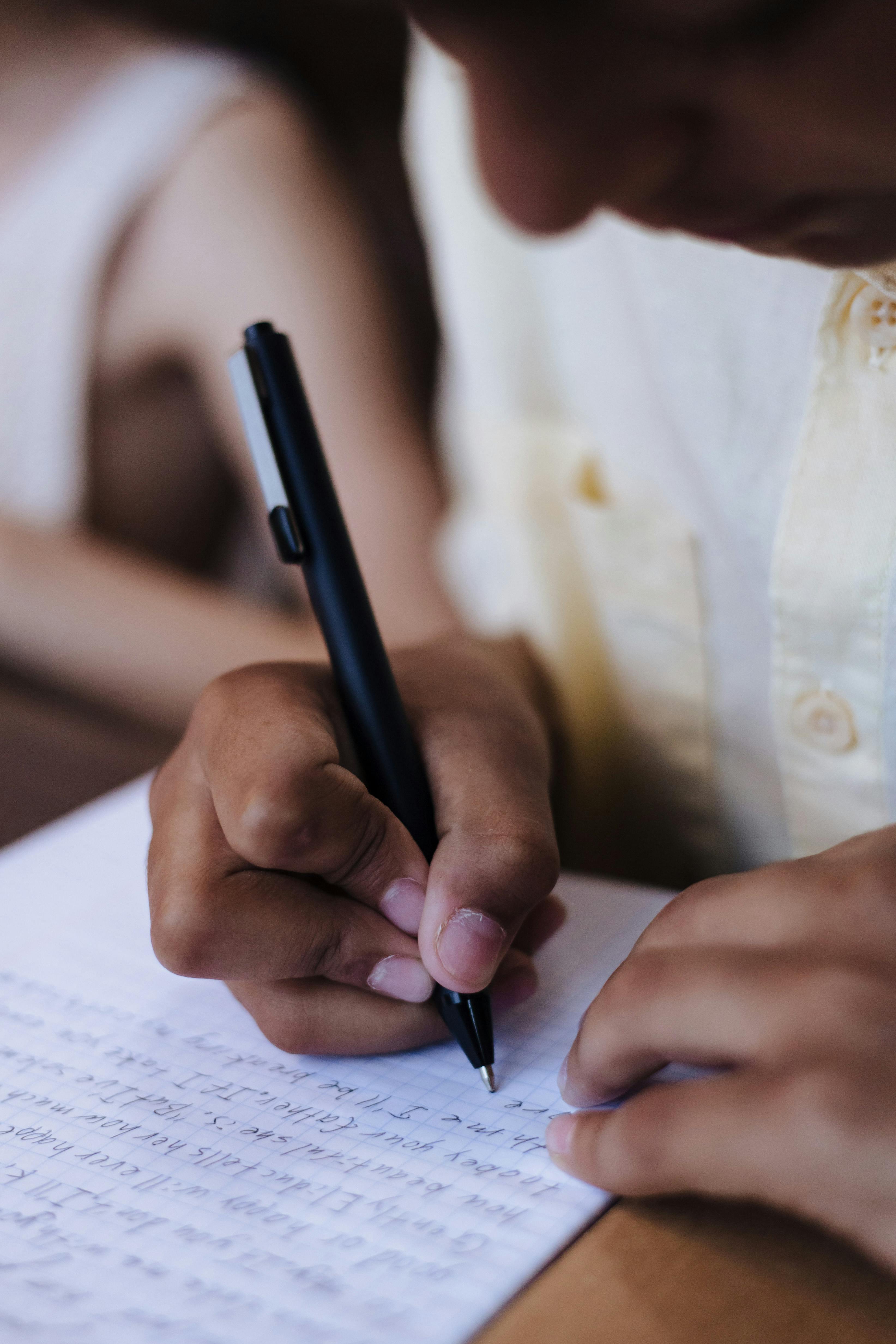 Child's hands writing with a pen on paper indoors, showing focus and concentration.